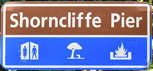 Brown sign for Shorncliffe Pier, blue sign with symbols for toilets, shaded picnic table, BBQ