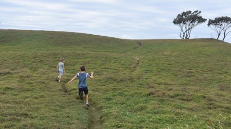 Two boys running on a grassy rolling hill with a couple of trees on the horizon, taken at Angourie Point in Yuraygir National Park