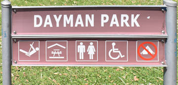 Brown sign for Dayman Park, symbols for swings, sheltered picnic tables, toilets, disabled access, no camping