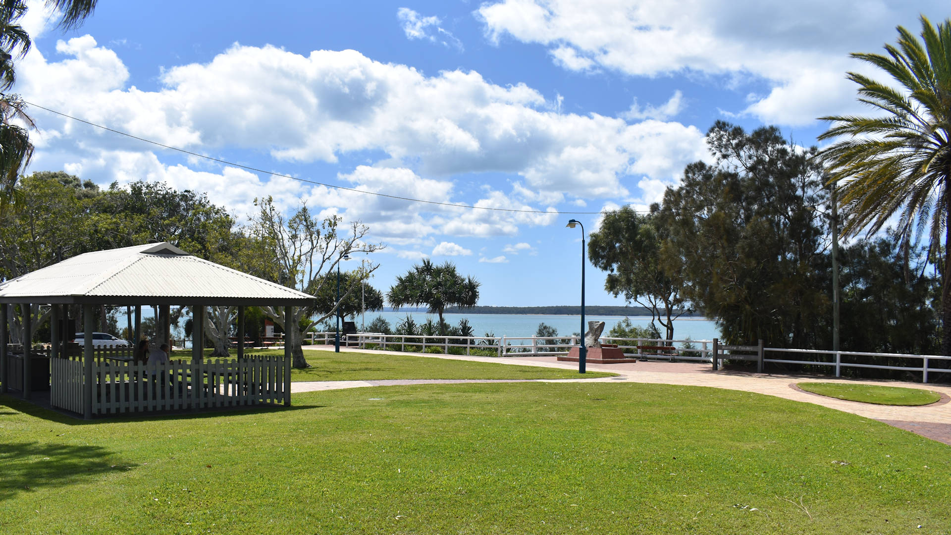Green grass area with a picnic table and BBQ shelter, bay water in the background, blue skies with scattered clouds, taken at Dayman Park in Hervey Bay