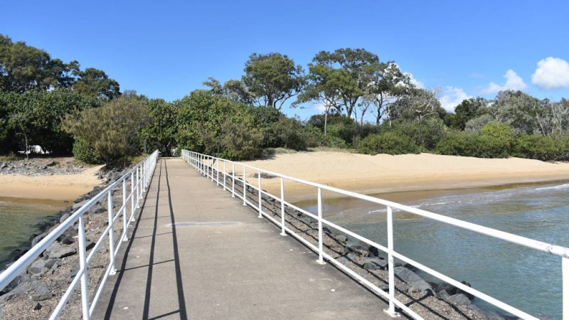 Looking towards land from a concrete jetty with metal railings on a rock wall, beach on both sides
