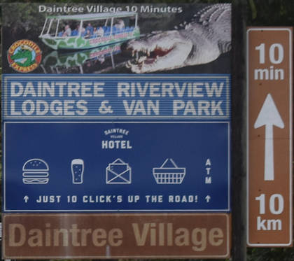 Brown sign for Daintree Village, vertical brown sign with up arrow, 10min and 10km, blue sign for Daintree Riverview Lodges & Van Park, symbols for food, drink, mail, groceries, ATM