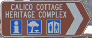 Brown sign for Calico Cottage Heritage Complex, blue symbols for visitor information, park, and toilets