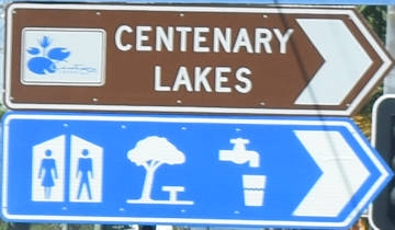Brown sign for Centenary Lakes, blue sign with symbols for toilets, shaded picnic tables, drinking water